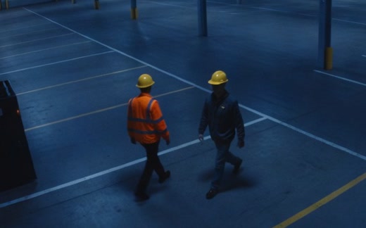 Two workers in hard hats in a warehouse seen from the ceiling. One wears a high-visibility vest, the other does not.